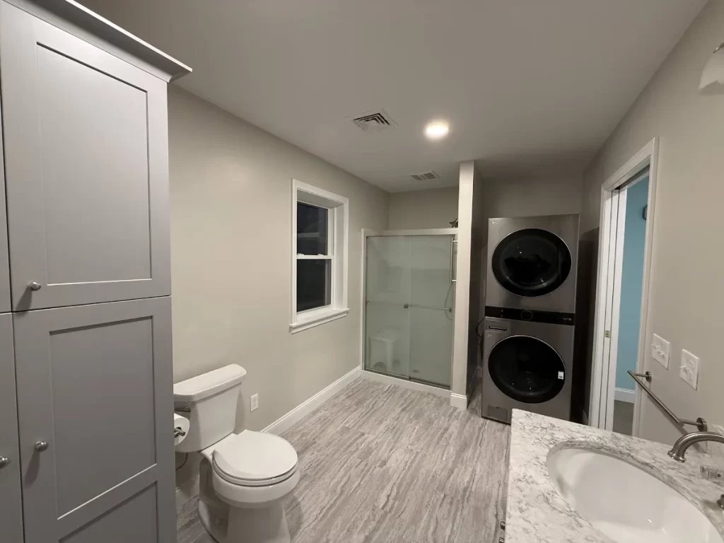 Remodeled bathroom with a glass walk-in shower, grab bar, stacked laundry, and gray cabinets by JB Cote Construction