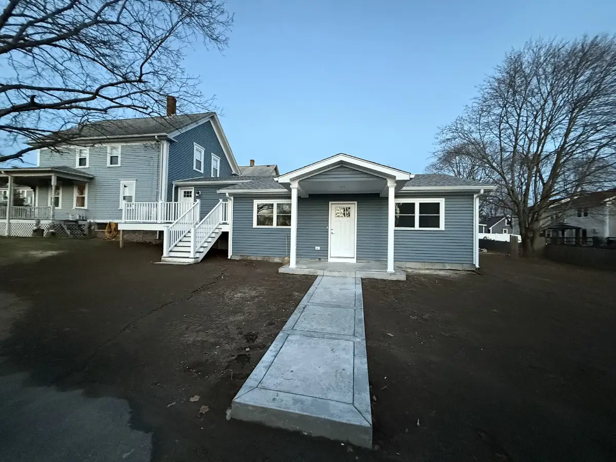 Single-story blue ADU with its own front door and concrete walkway behind a Rhode Island home, built by JB Cote Construction